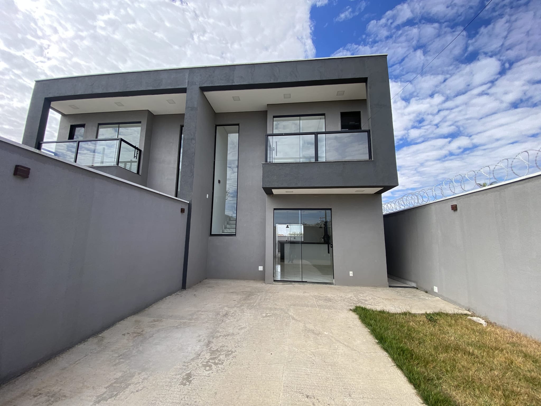Fachada da residência vista do recuo, com dois pavimentos, varanda com guarda-corpo de vidro e acesso em porcelanato.